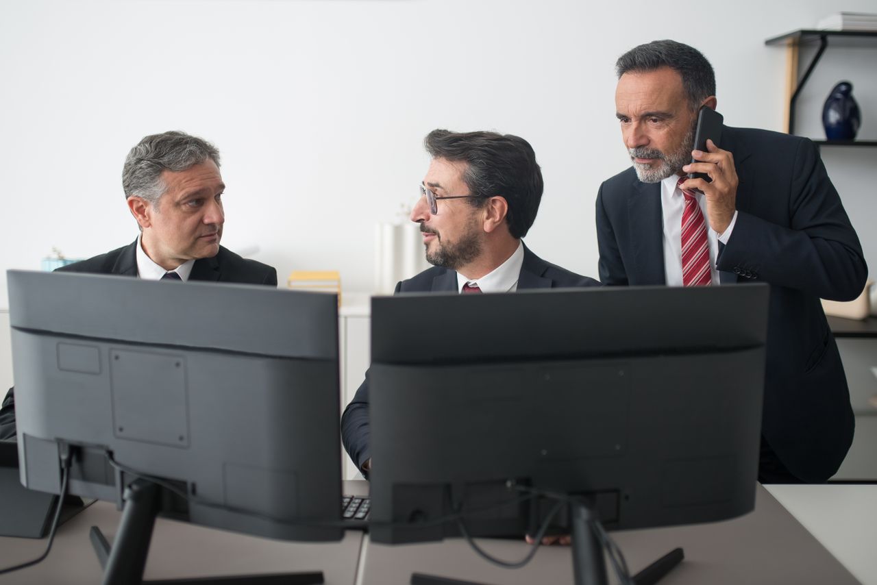 Crafting Captivating Headlines: Your awesome post title goes here Three businessmen in an office discussing work, two using computers and one on a phone call.